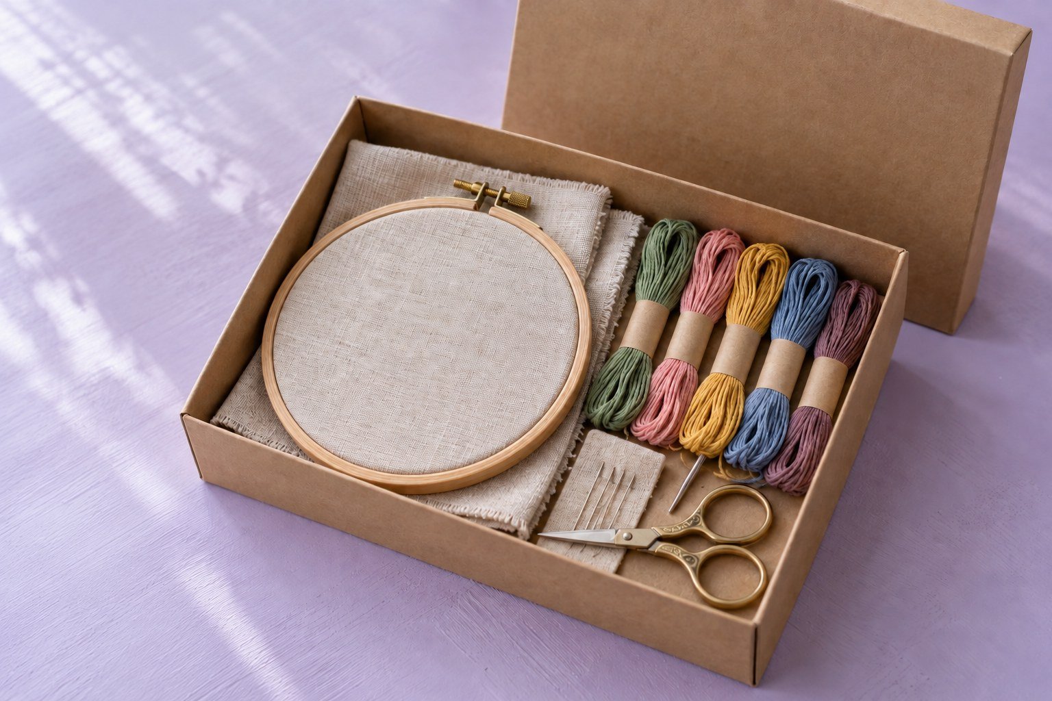 An open gift box containing an embroidery hoop, threads and linen